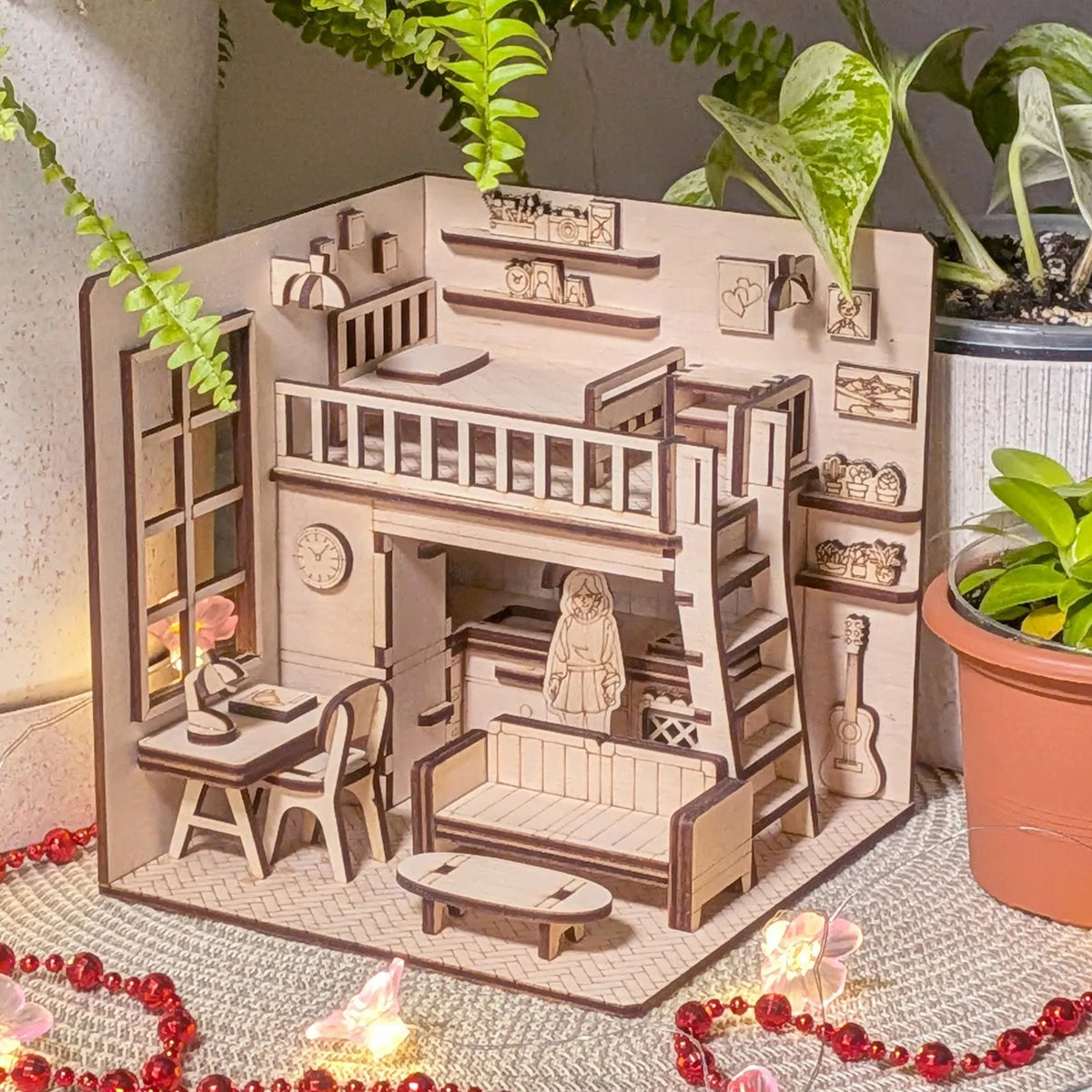 Wooden model of a house with furniture and decorative items, surrounded by plants and red beads.