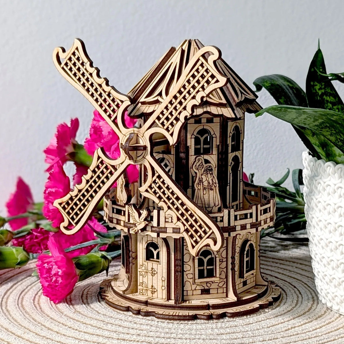Decorative 3D plywood windmill model with ornate architectural details, photographed on a woven table mat next to indoor plants.