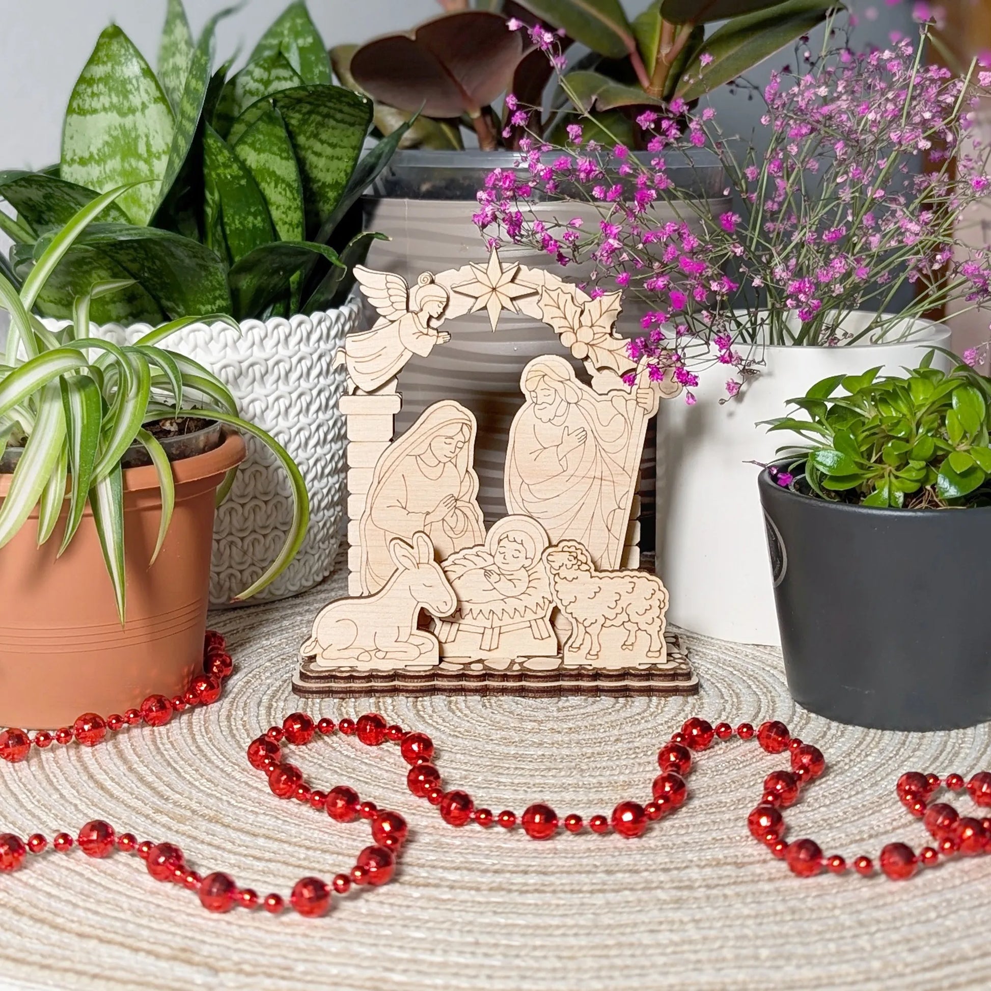 Wooden nativity scene with potted plants and red beads on a textured surface