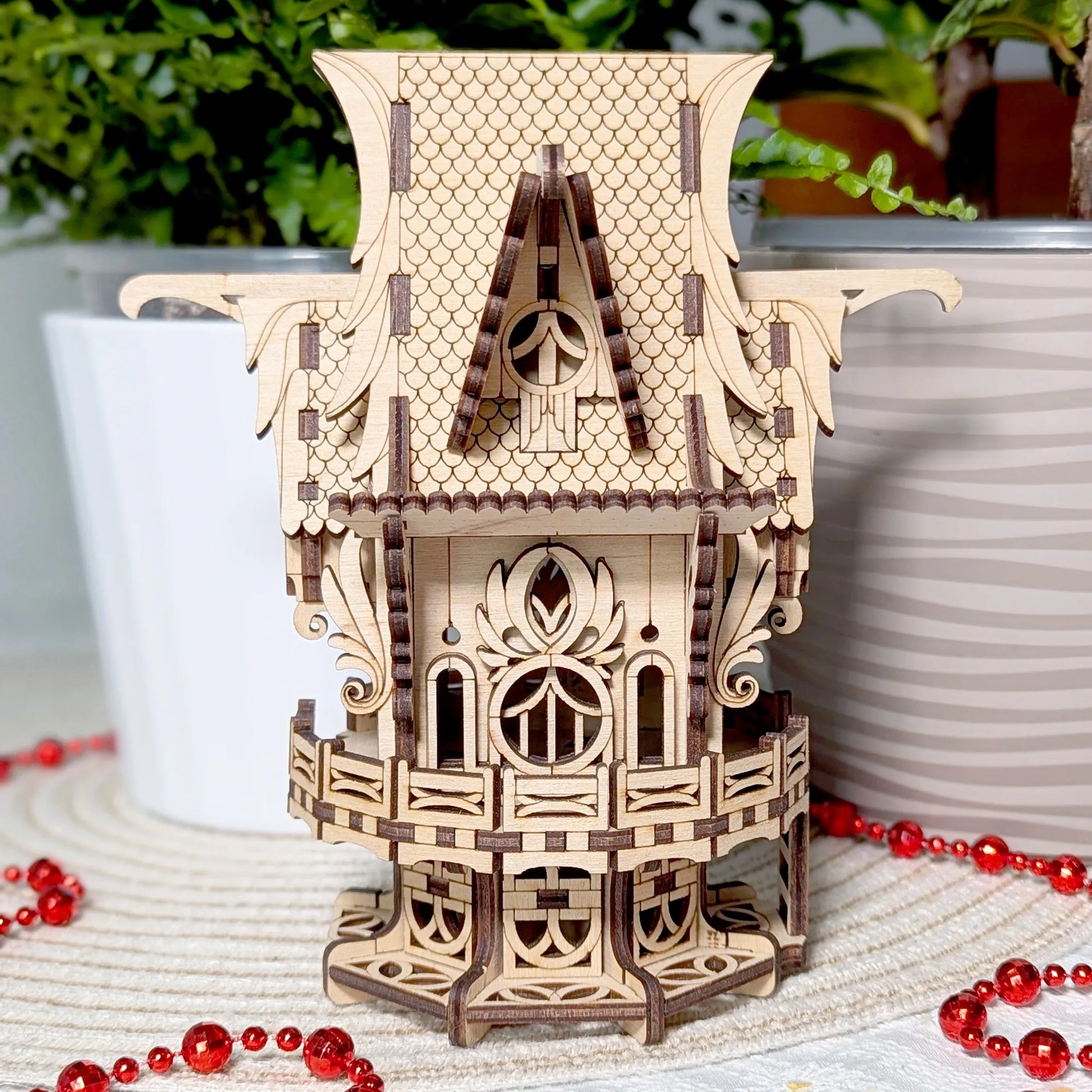 Wooden birdhouse model of a with decorative elements on a surface with red beads and plants in the background.