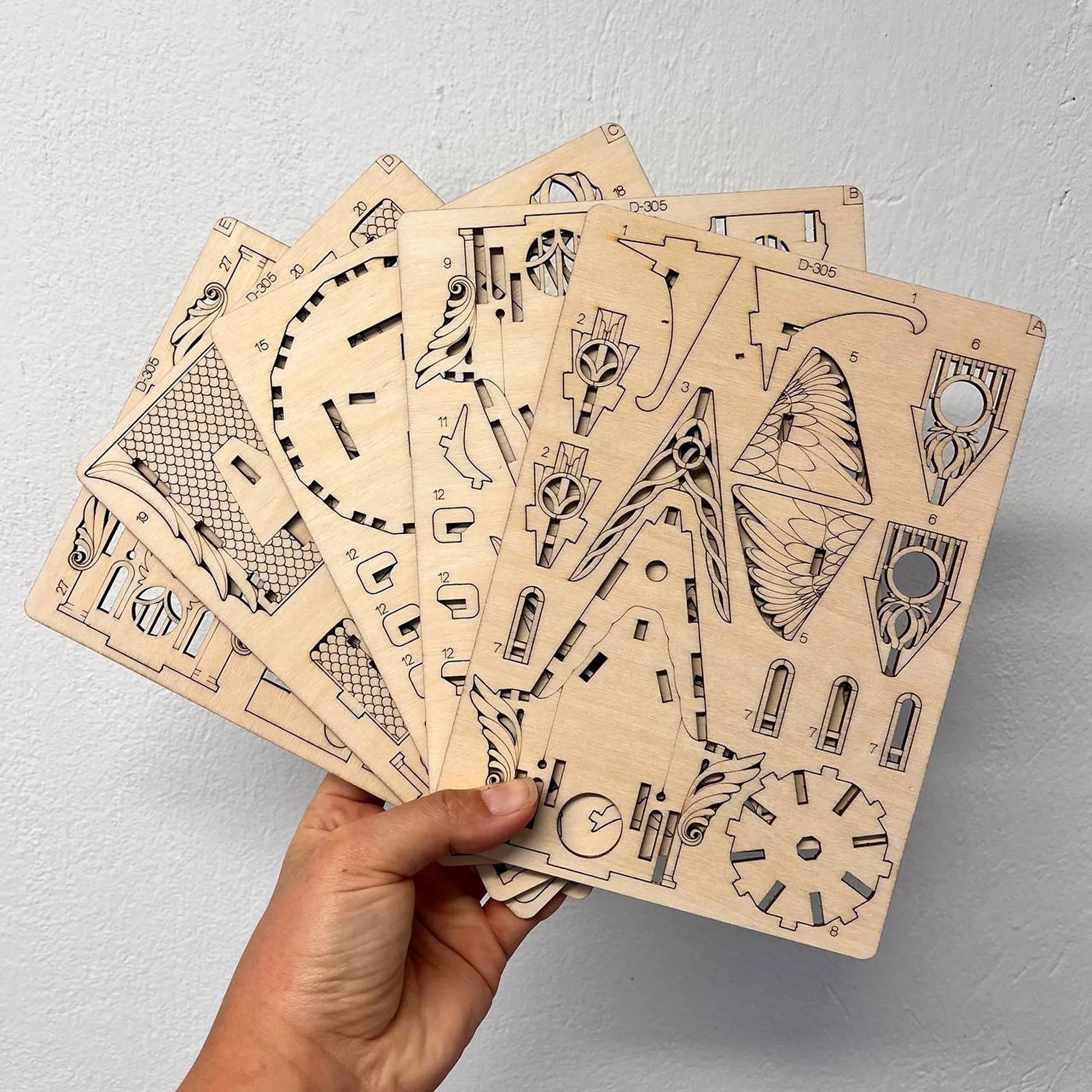 A hand holds a fan of wooden craft panels with intricate designs, including a clock, gears, and wing-like shapes, against a light gray textured wall.