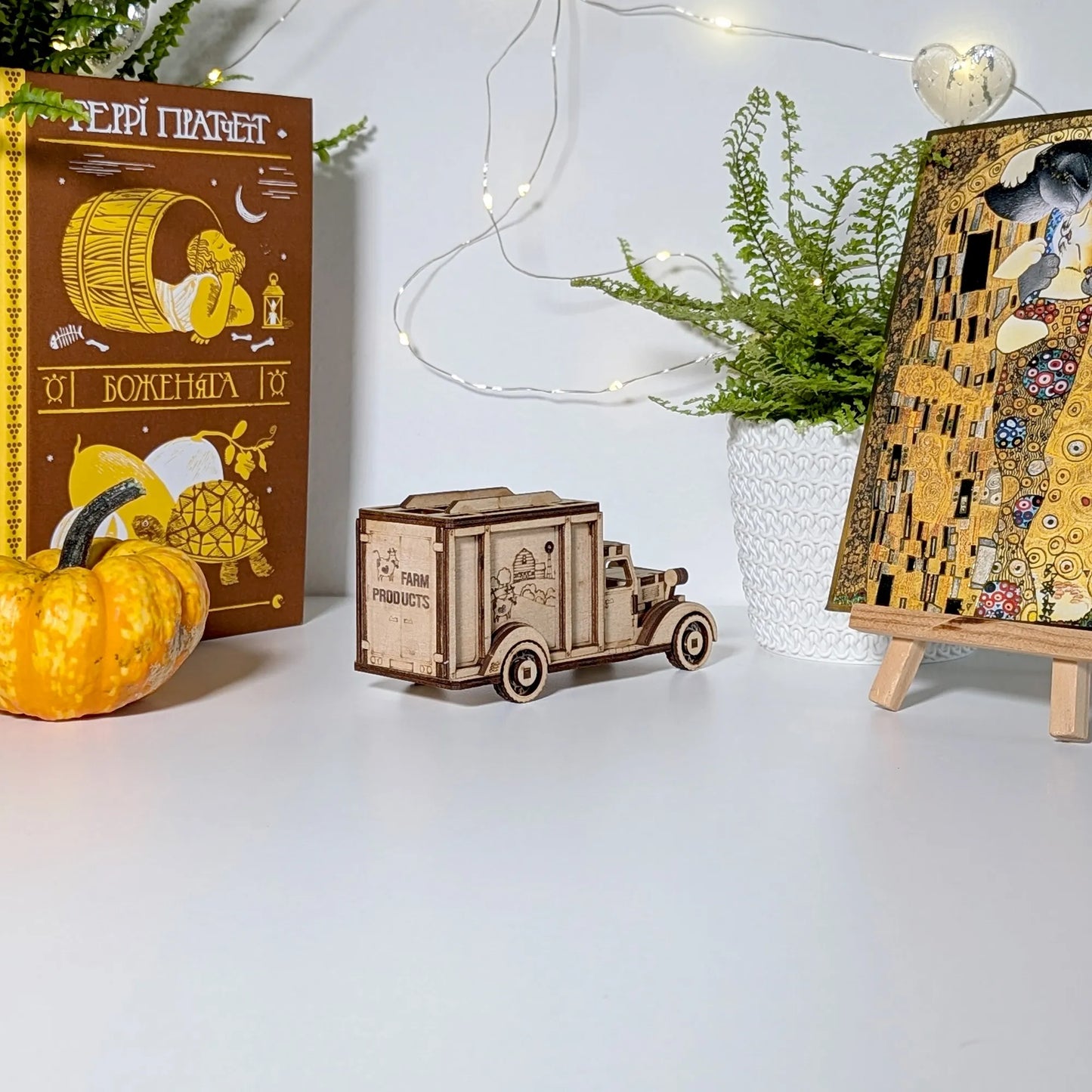 Wooden toy truck, pumpkin, and framed artwork on a white surface with decorative lights and plants.