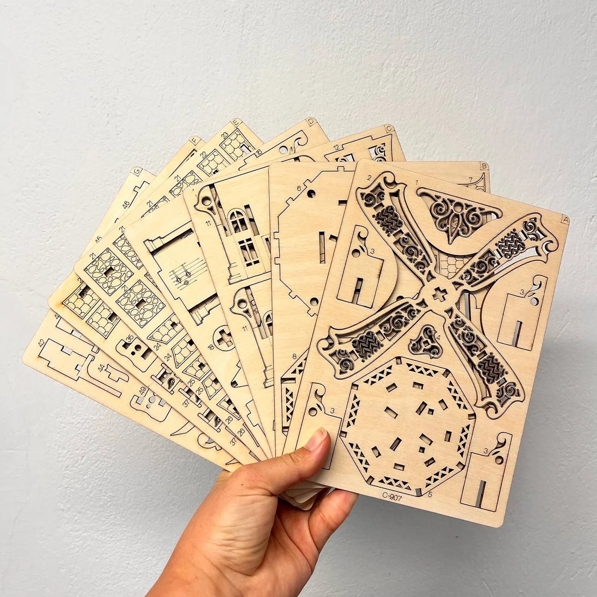 A hand holds a fanned-out set of wooden craft templates with intricate architectural and decorative designs, shown against a plain light gray wall.