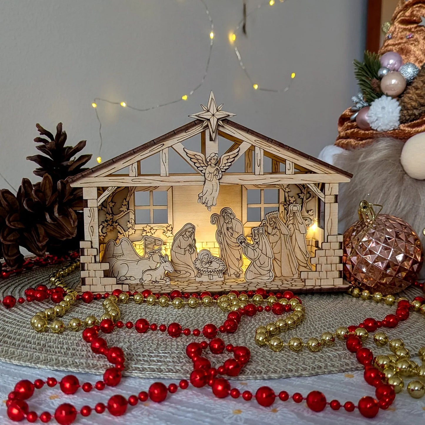 Wooden nativity scene with lights surrounded by red and gold beads on a textured surface.