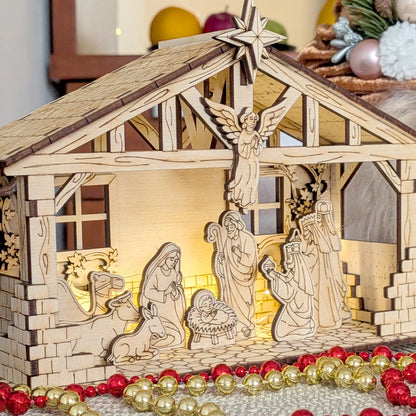 Wooden nativity scene model with illuminated interior, surrounded by Christmas decorations.