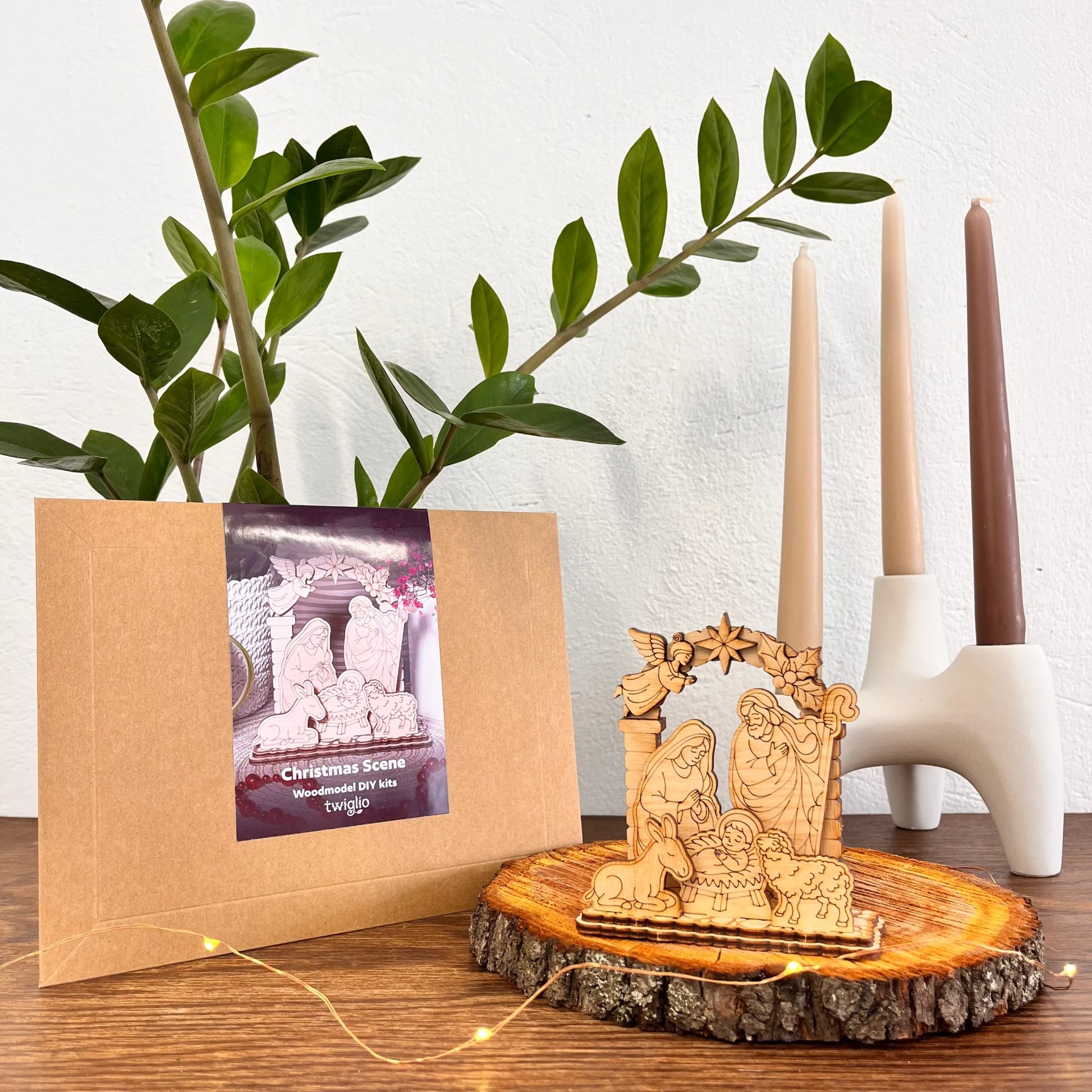 Wooden model Christmas scene on a wooden stand with a plant and candle holder in the background.