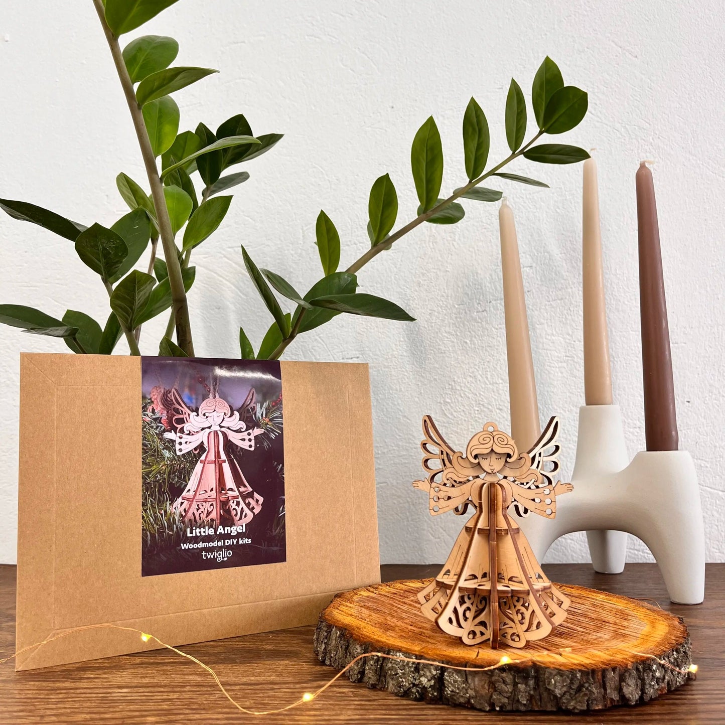 Wooden model angel on a wooden stand with a plant and candle holder in the background.