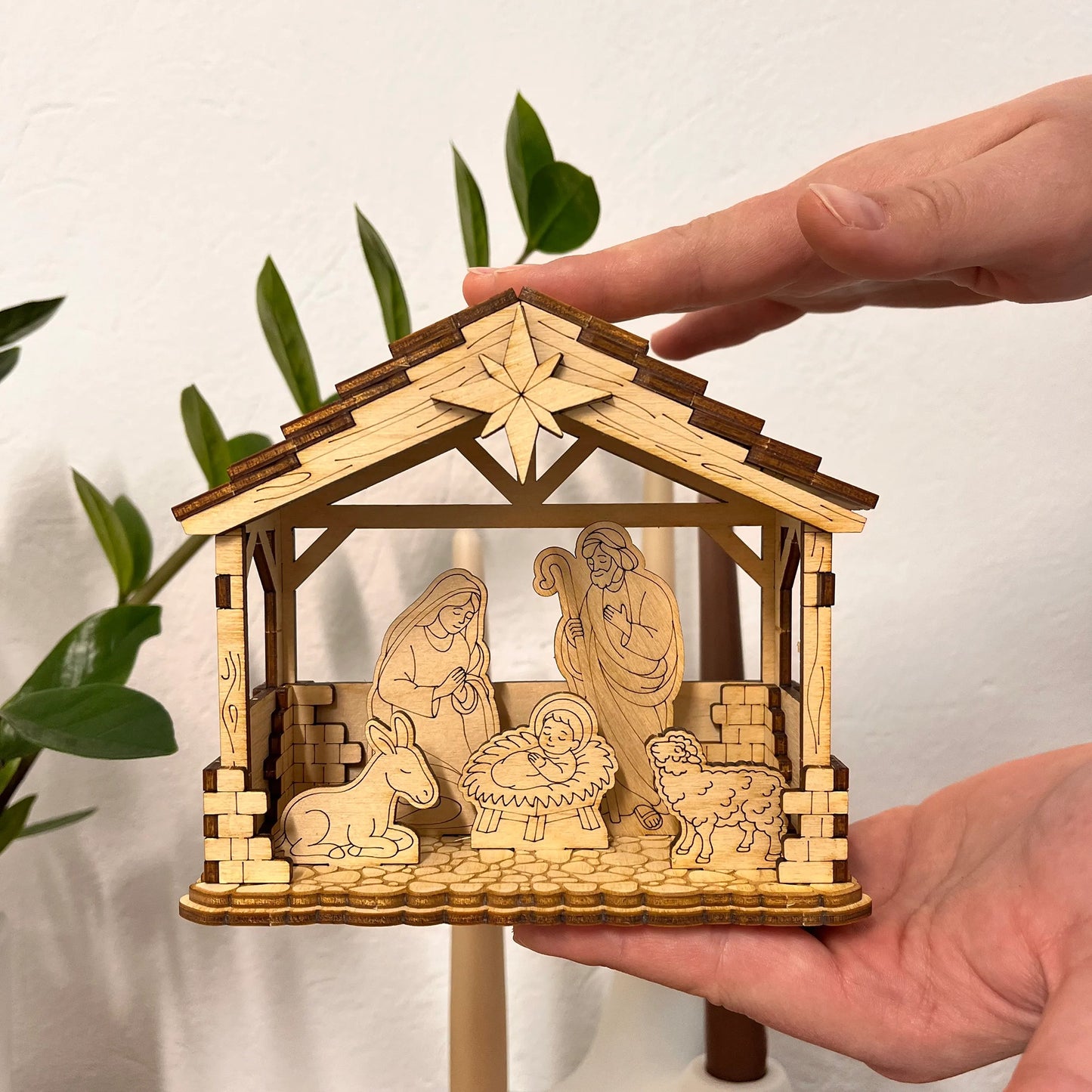 Wooden nativity scene model held by a hand with a plant in the background
