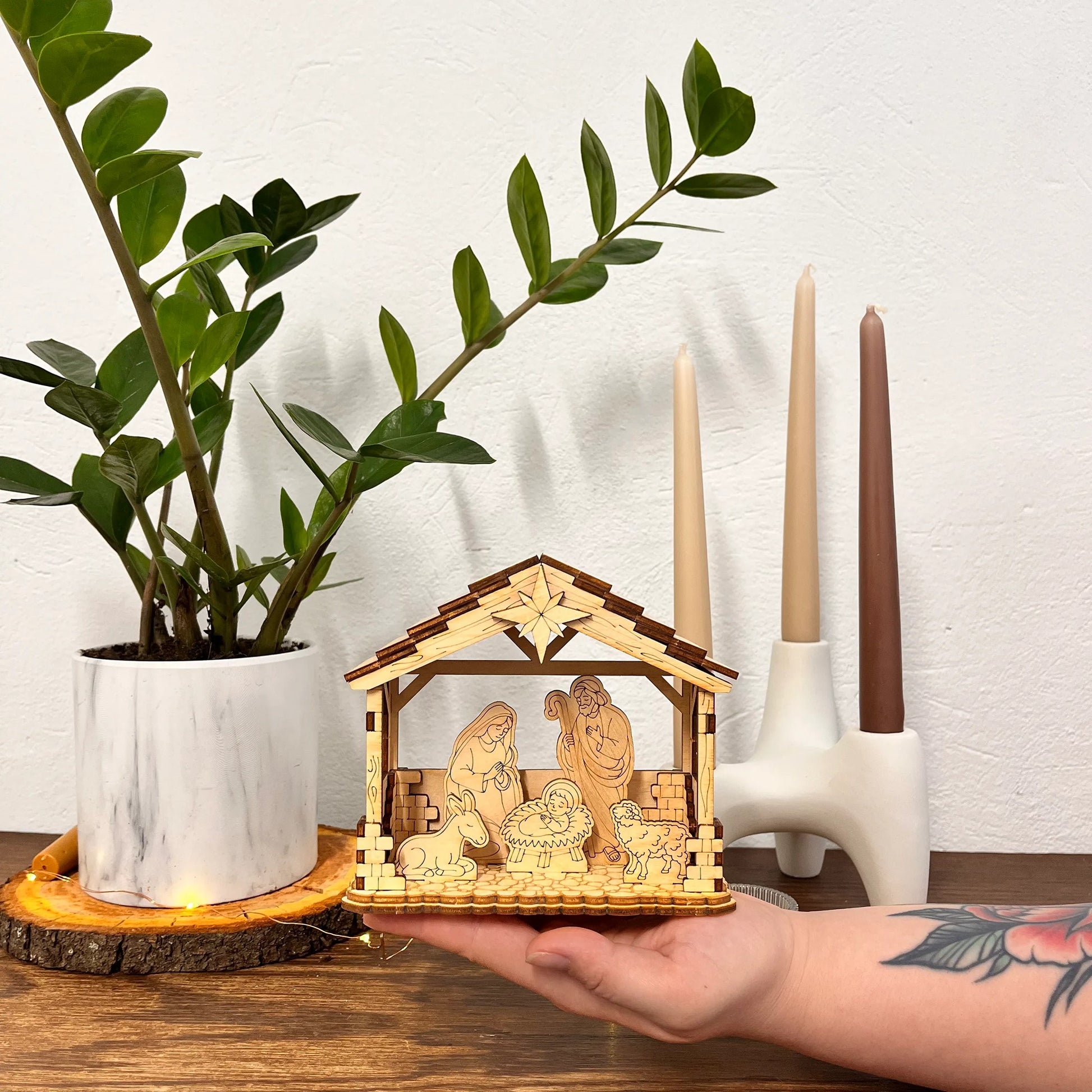 Hand holding a small nativity scene model with candles and a plant in the background.