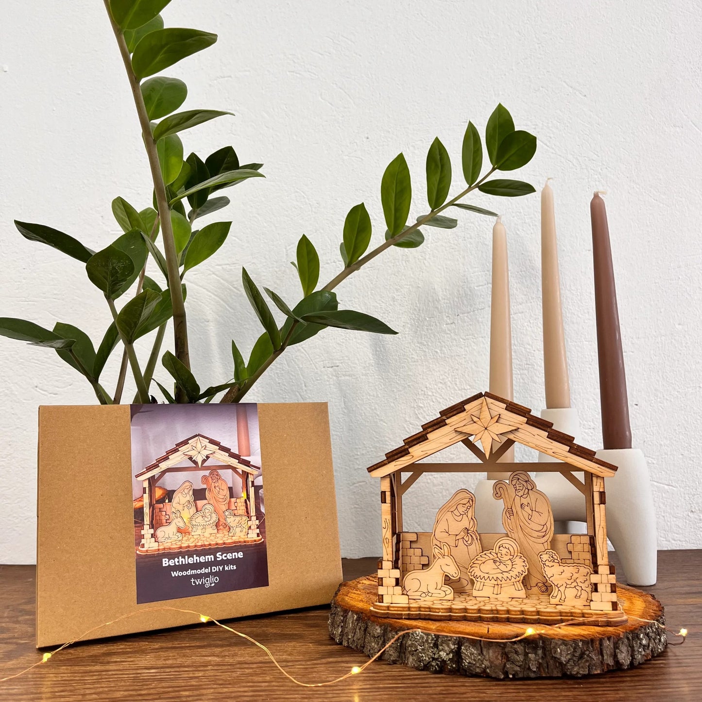 Wooden nativity scene with a cardboard packaging box featuring an illustration of a nativity scene, placed on a wooden surface with candles and a plant in the background.