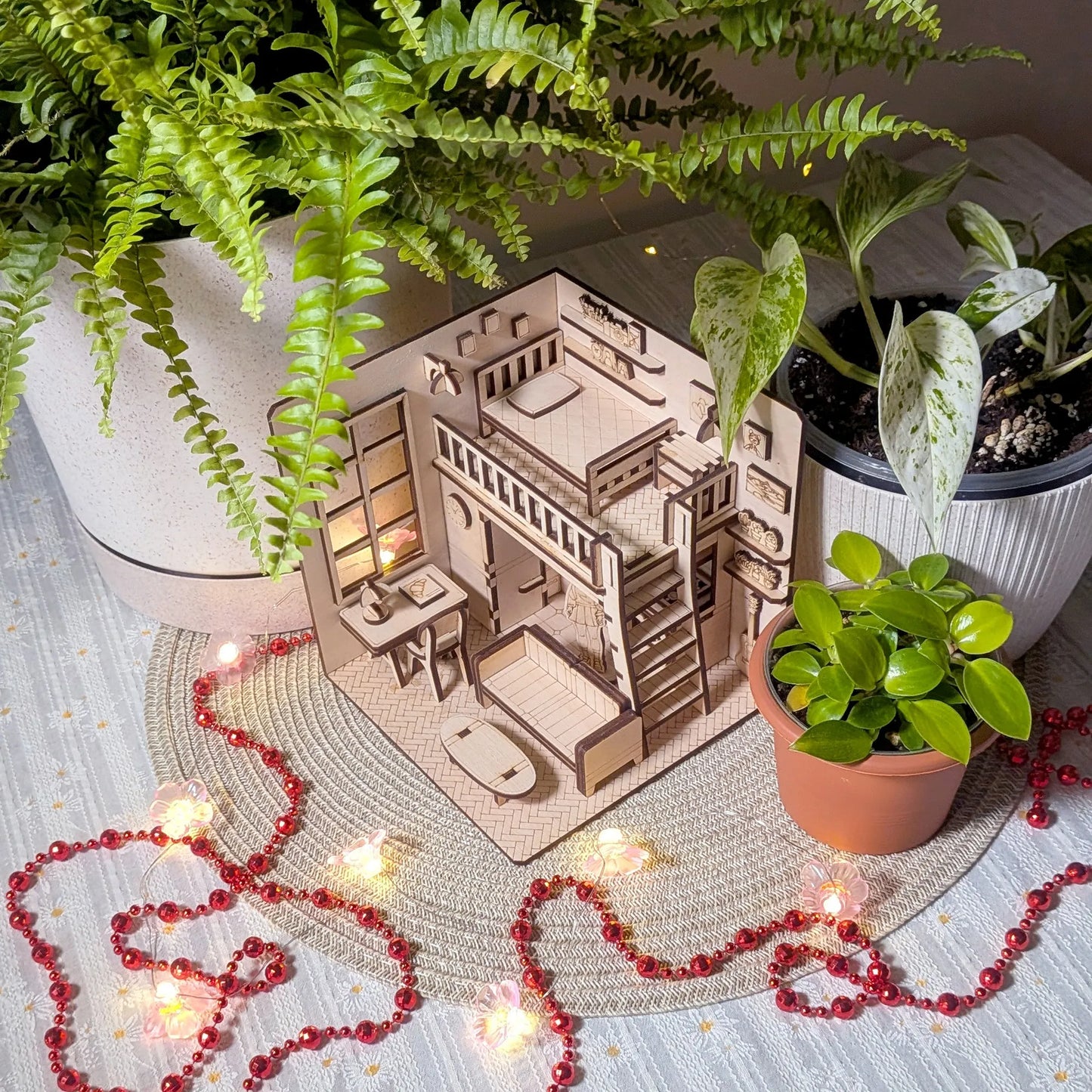 Miniature wooden house model with furniture surrounded by potted plants on a textured surface.