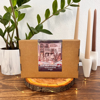 Wooden model kit packaging with a book nook greenhouse on a wooden surface with plants and candles in the background.