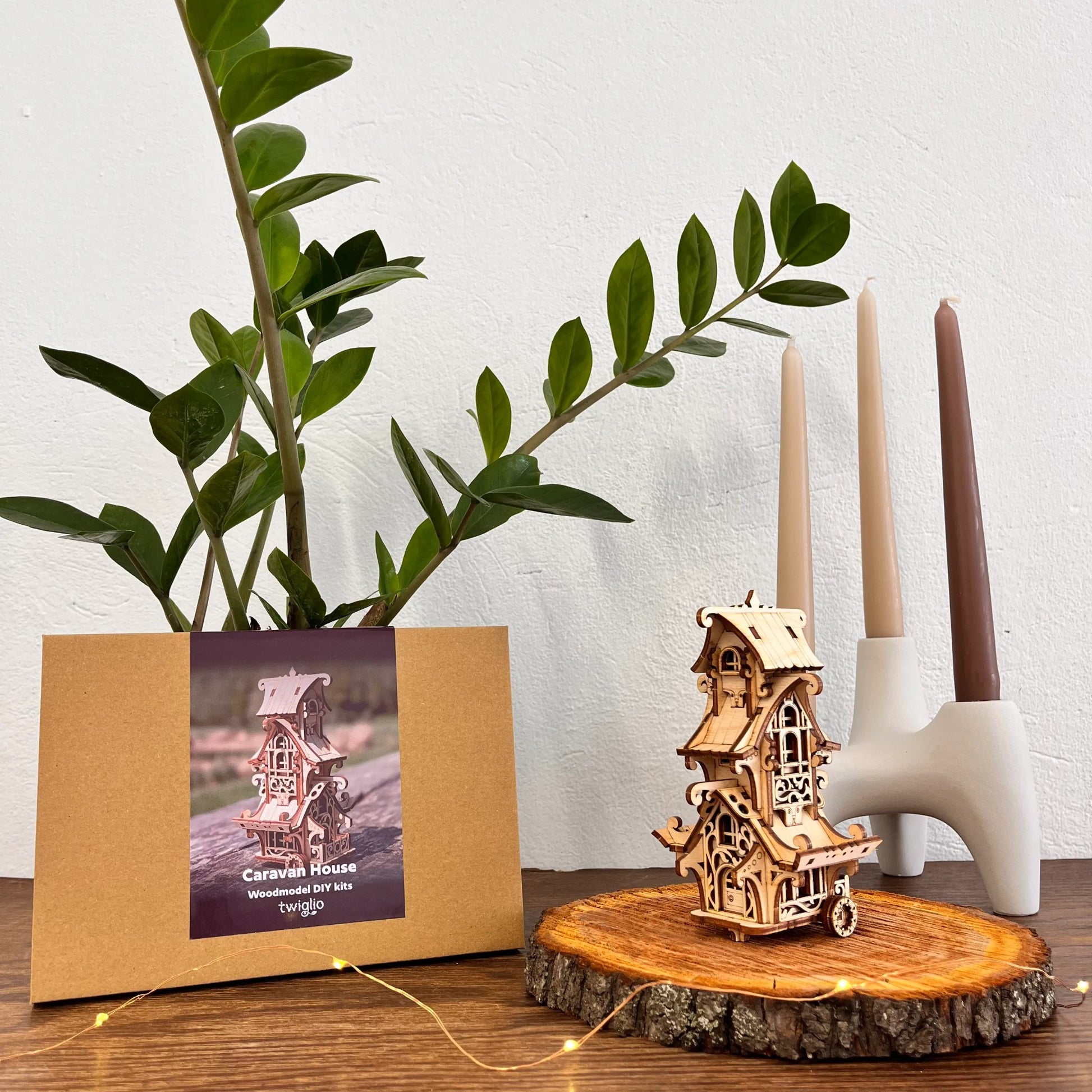 Wooden model caravan house on a wooden stand with a plant and candle holder in the background.