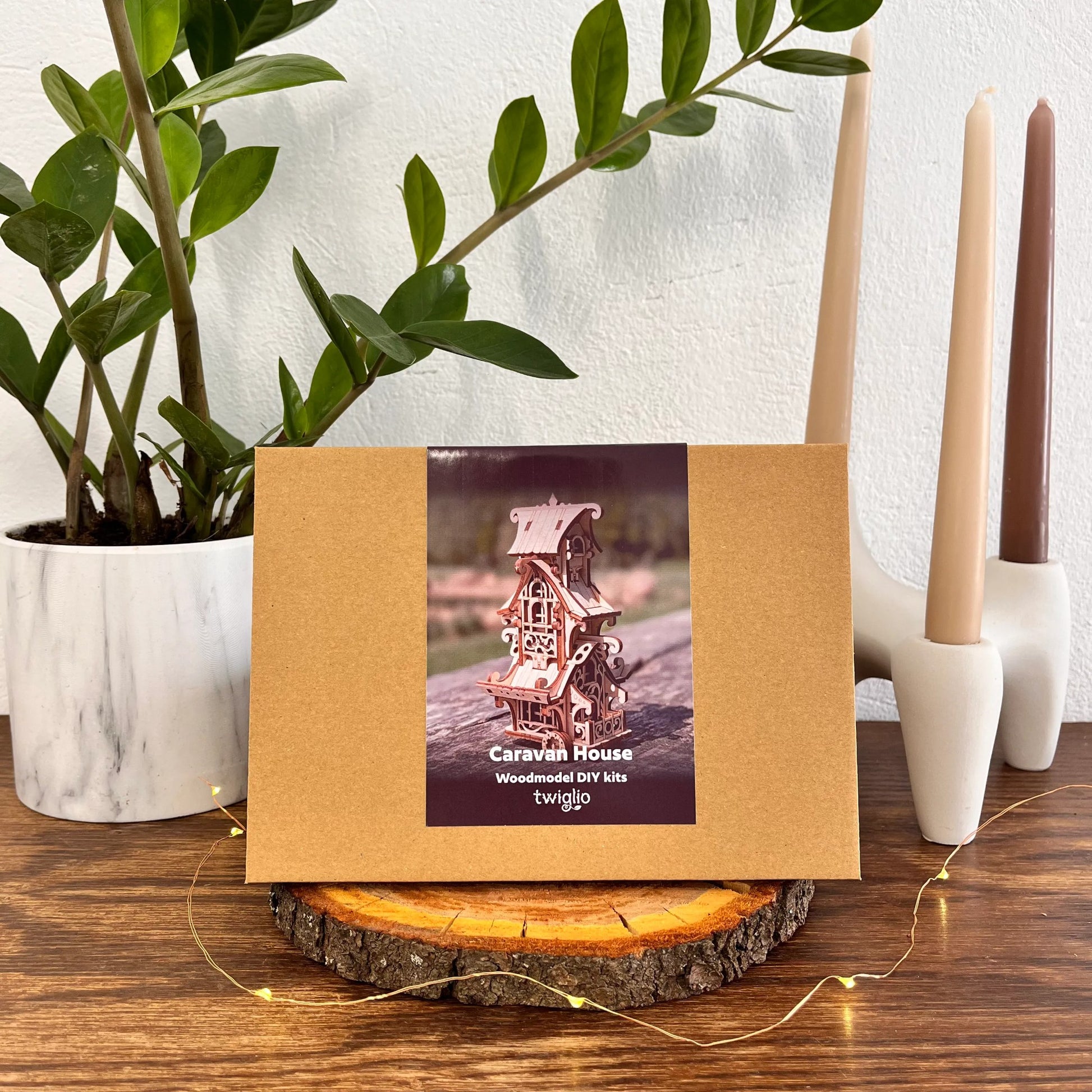 Wooden model kit packaging with a miniature caravans house on a wooden surface with plants and candles in the background.