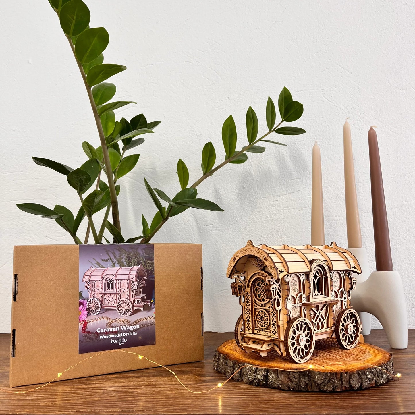 Wooden model of a caravan on a wooden stand with a plant and candles in the background.