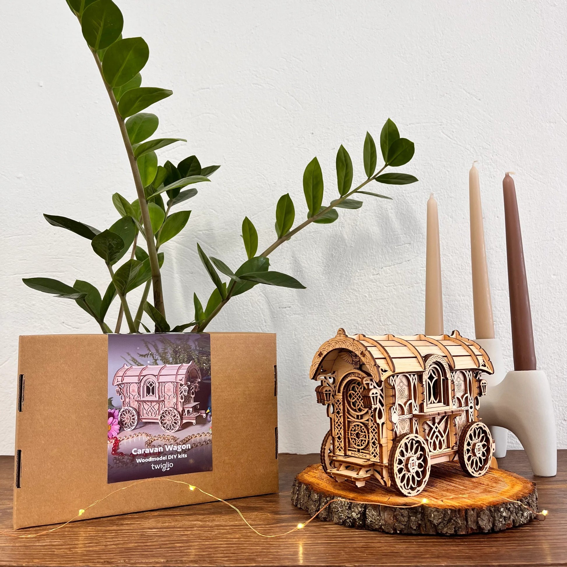 Wooden model of a caravan on a wooden stand with a plant and candles in the background.