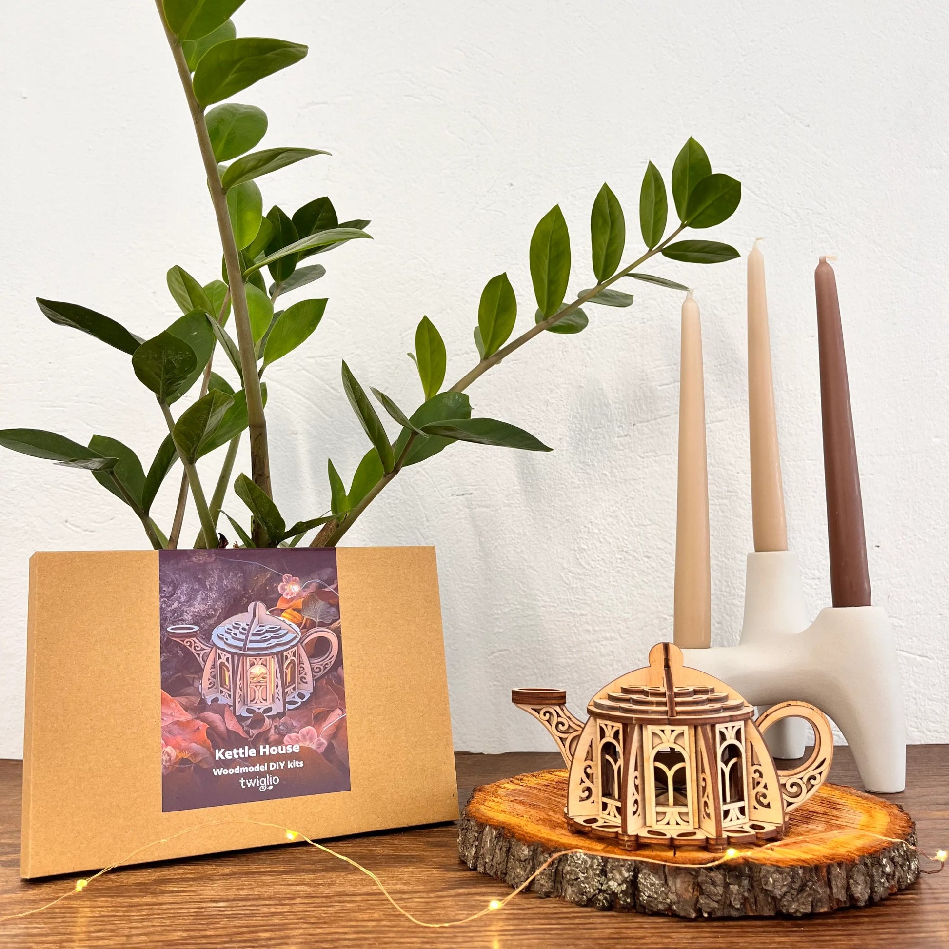 Wooden teapot model on a wooden stand with candles and a plant in the background