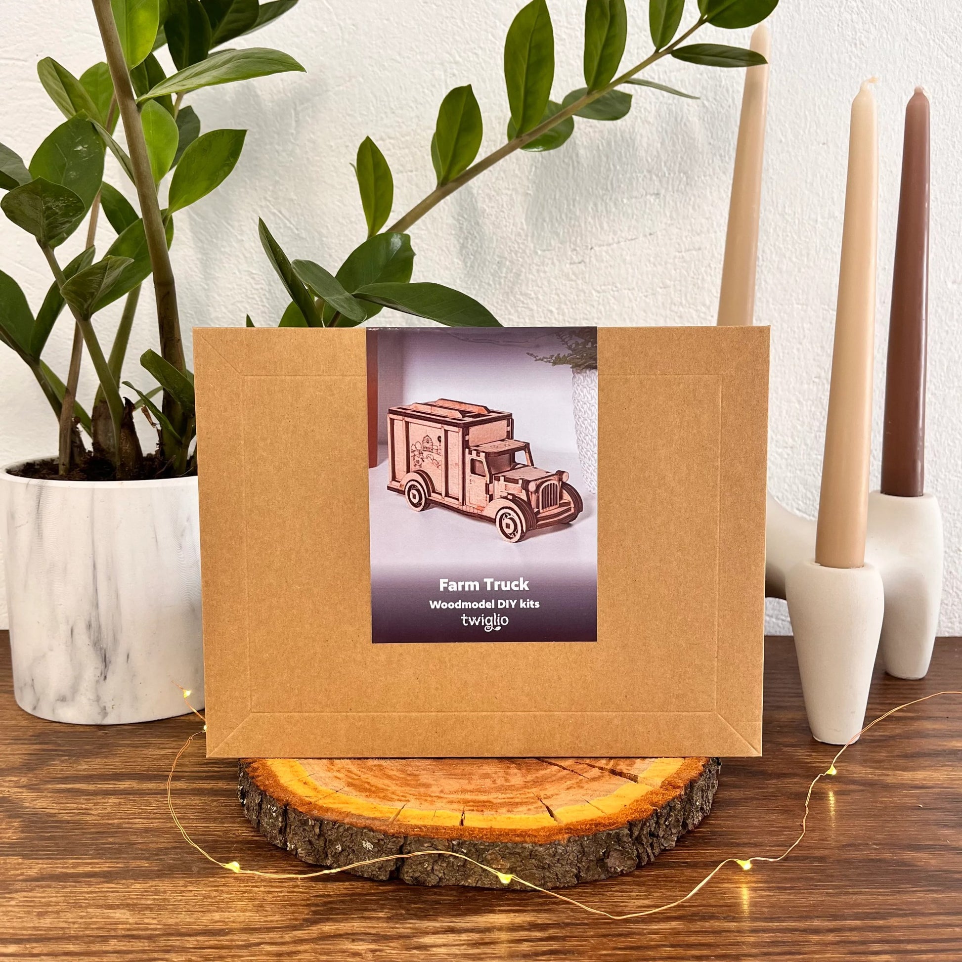 Wooden model kit of a farm truck in a brown box on a wooden surface with plants and candles in the background.