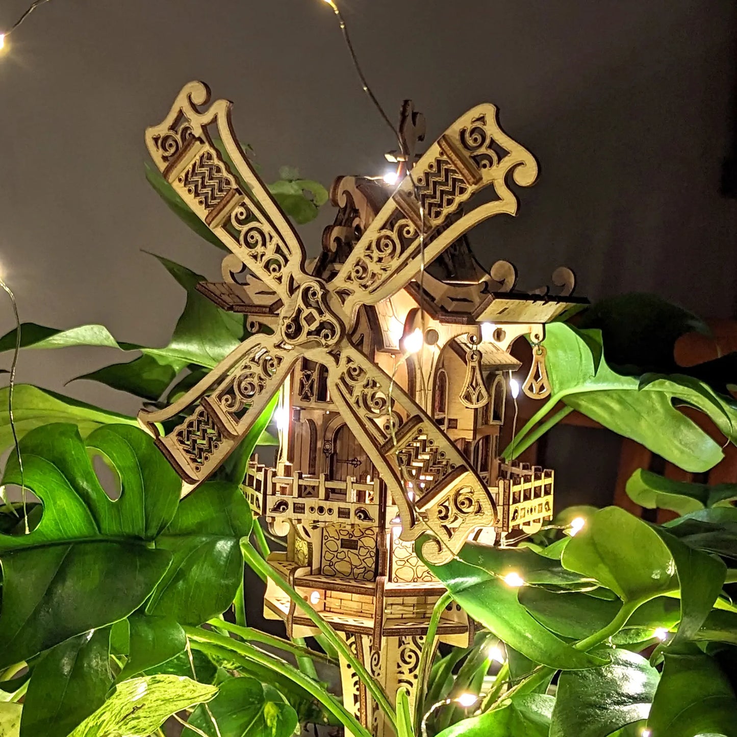 Decorative wooden windmill with intricate details, surrounded by green leaves and string lights.