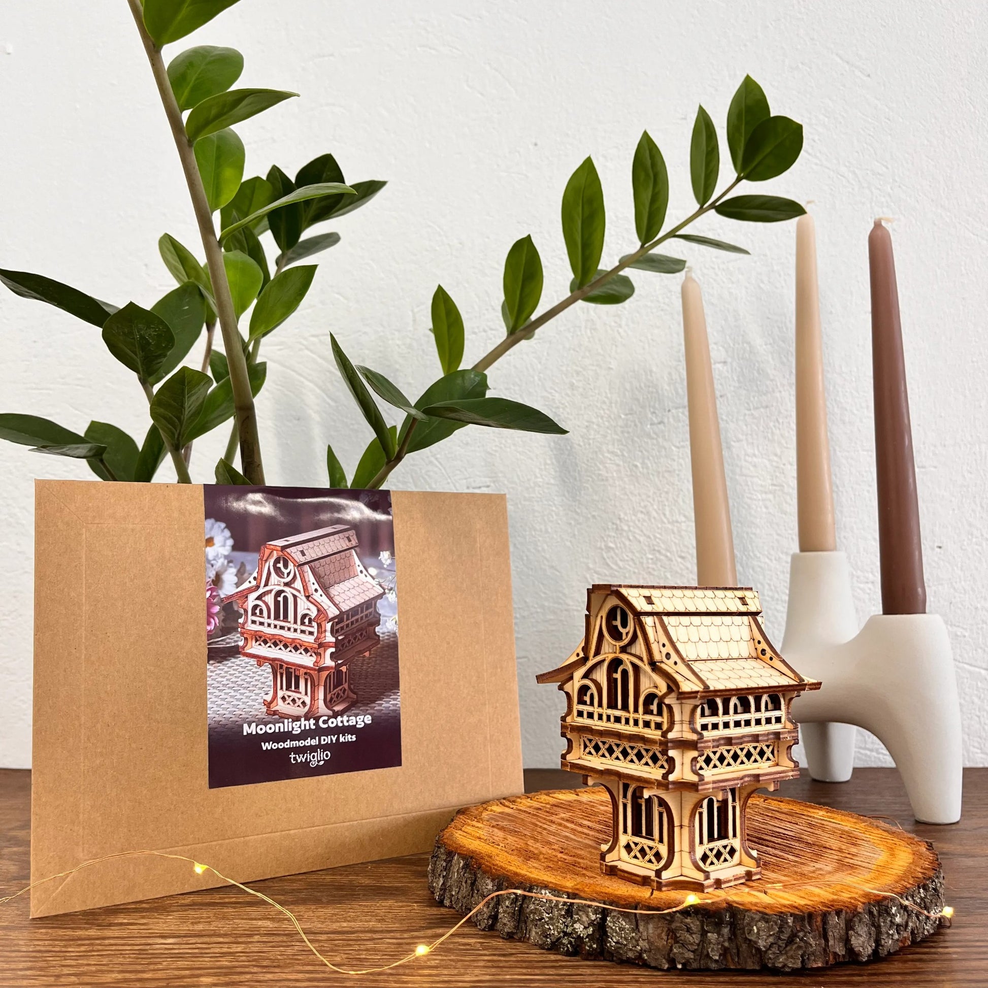 wooden DIY kit packaging with cottage model on a wooden surface with plants and candles in the background.