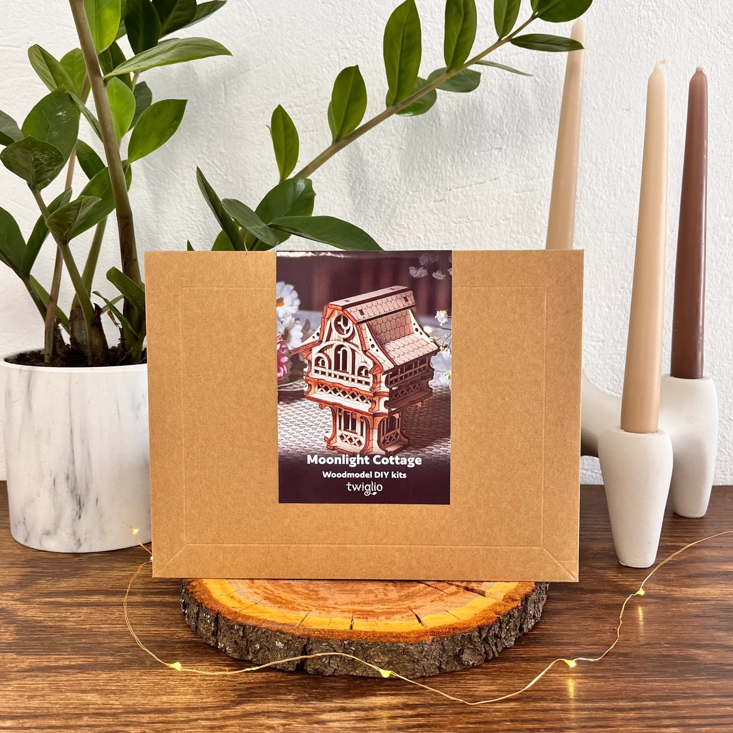 wooden DIY kit packaging of cottage model on a wooden surface with plants and candles in the background.