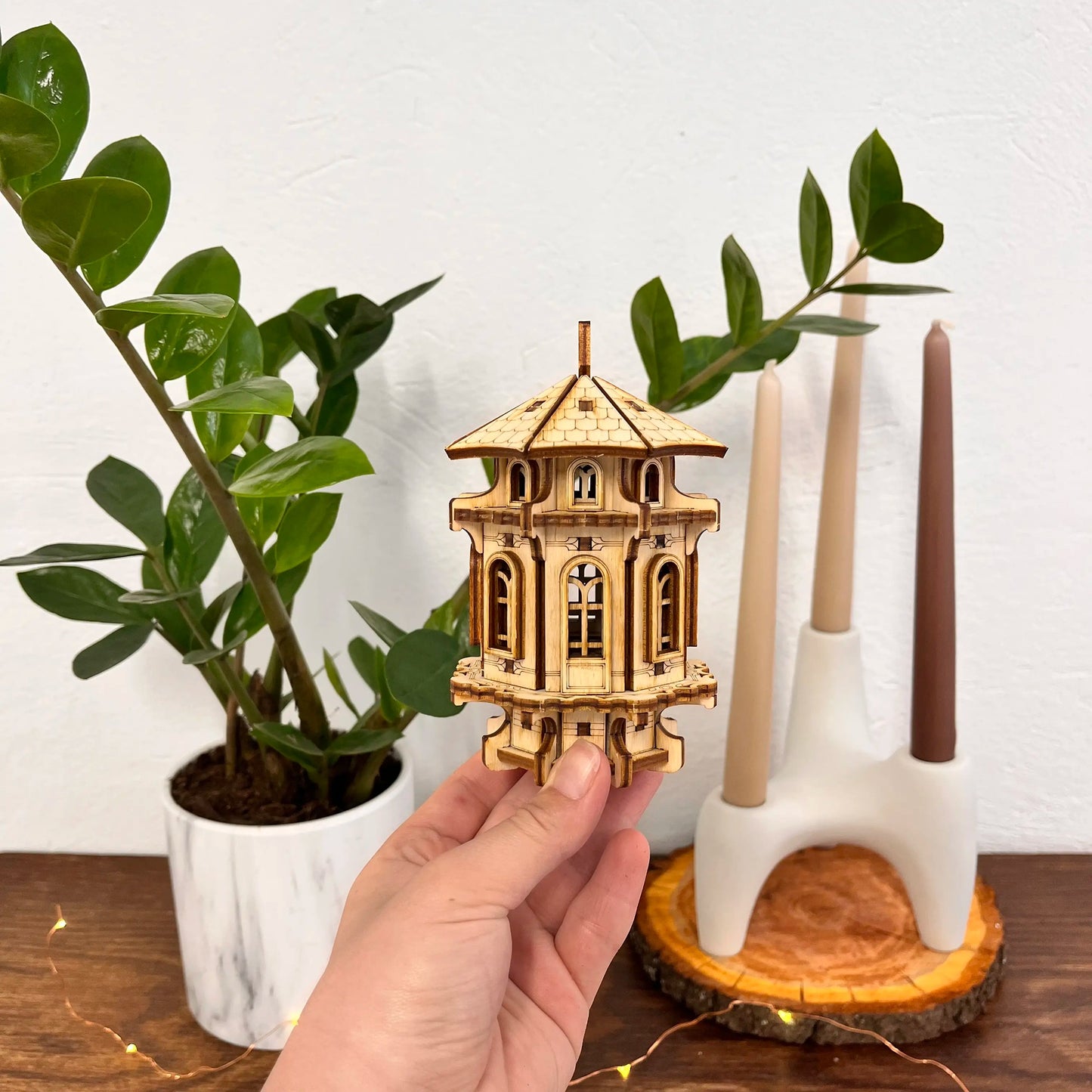 Wooden model of a pagoda on a hand with a plant and candle holder in the background.