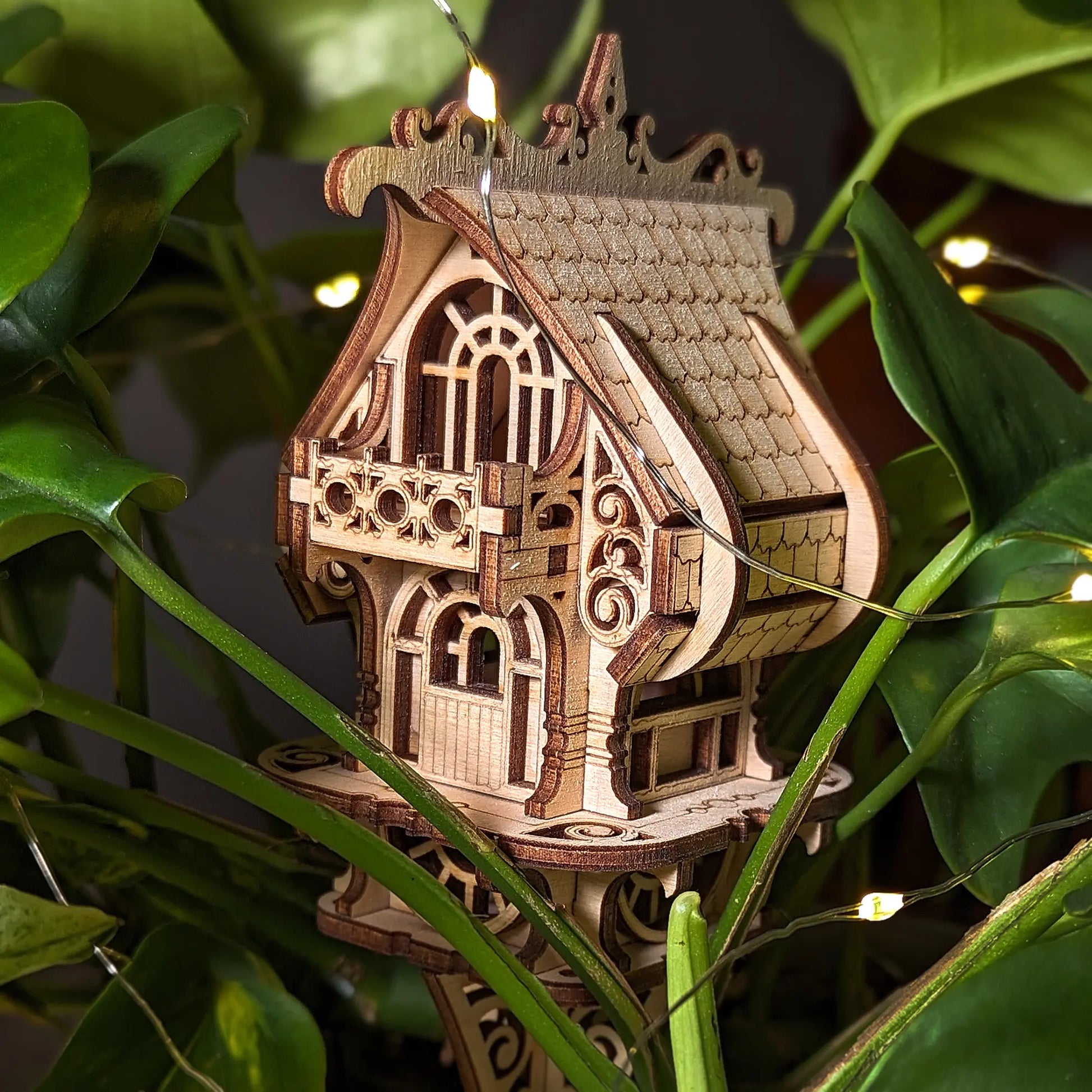 Wooden house ornament with intricate details hanging among green leaves.