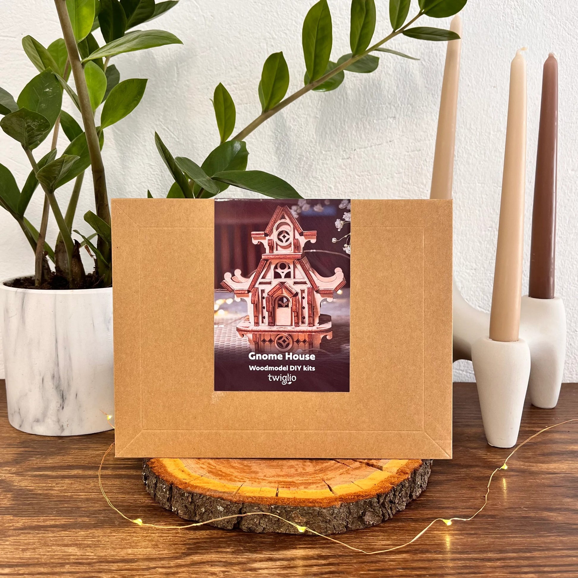 Wooden DIY kit packaging of garden gnome house on a wooden surface with plants and candles in the background.