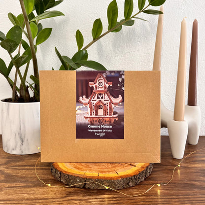 Wooden DIY kit packaging of garden gnome house on a wooden surface with plants and candles in the background.