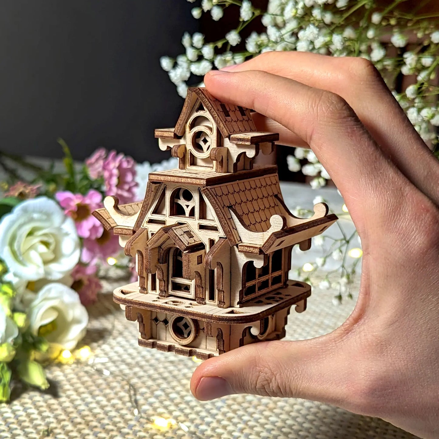 Hand holding a wooden model of a pagoda-like structure with flowers in the background