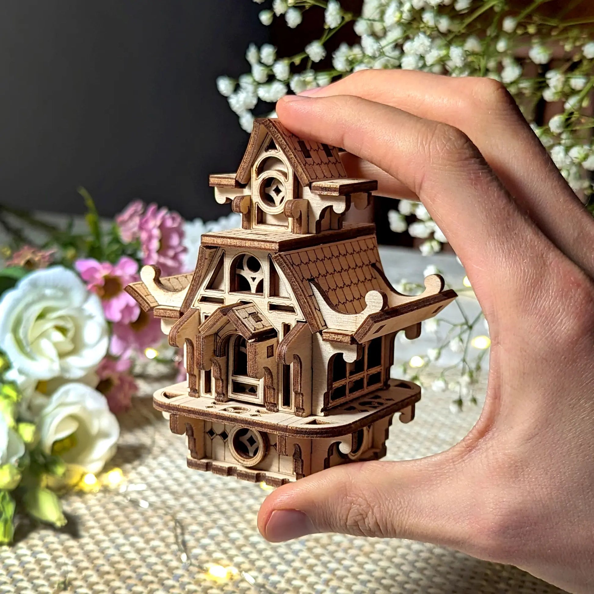 Hand holding a wooden model of a pagoda-like structure with flowers in the background