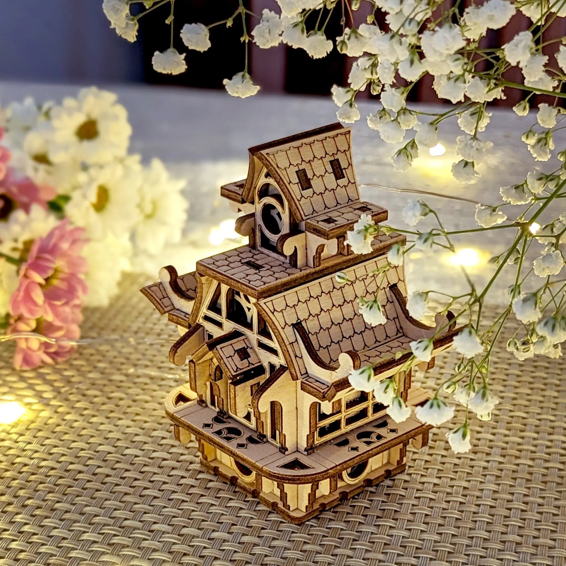 Wooden model of a traditional building with flowers and string lights on a woven surface