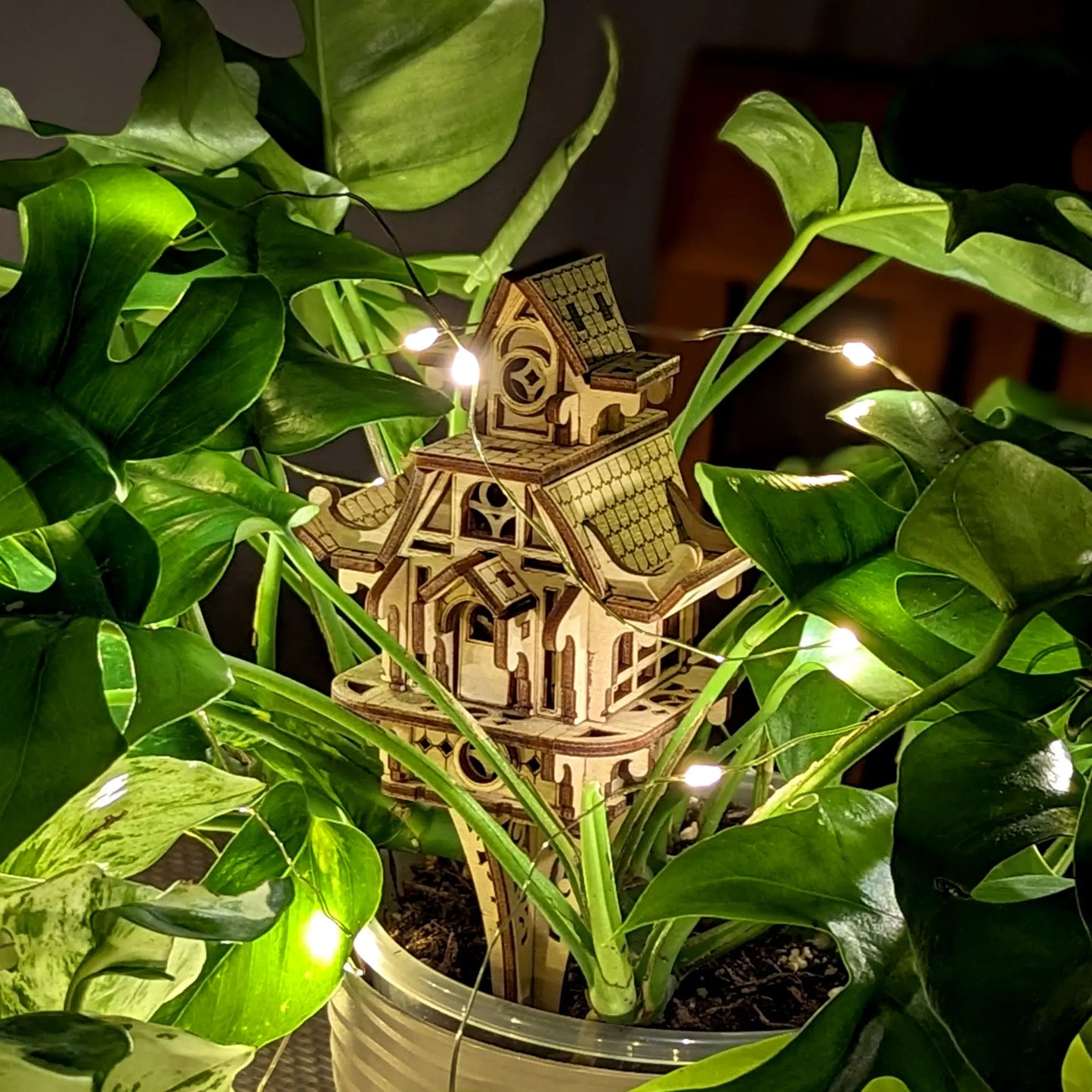 Wooden model of a house surrounded by green plants with string lights.