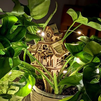 Wooden model of a house surrounded by green plants with string lights.