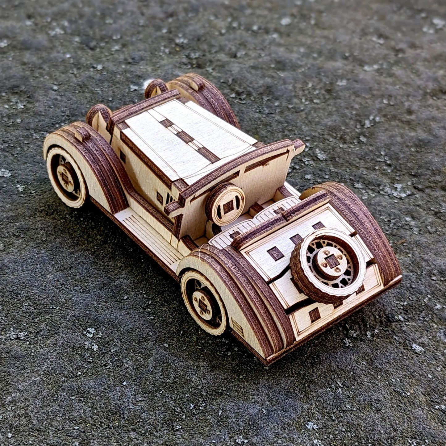 Vintage-style wooden sports car miniature made from laser-cut plywood, displayed on a rough outdoor surface.