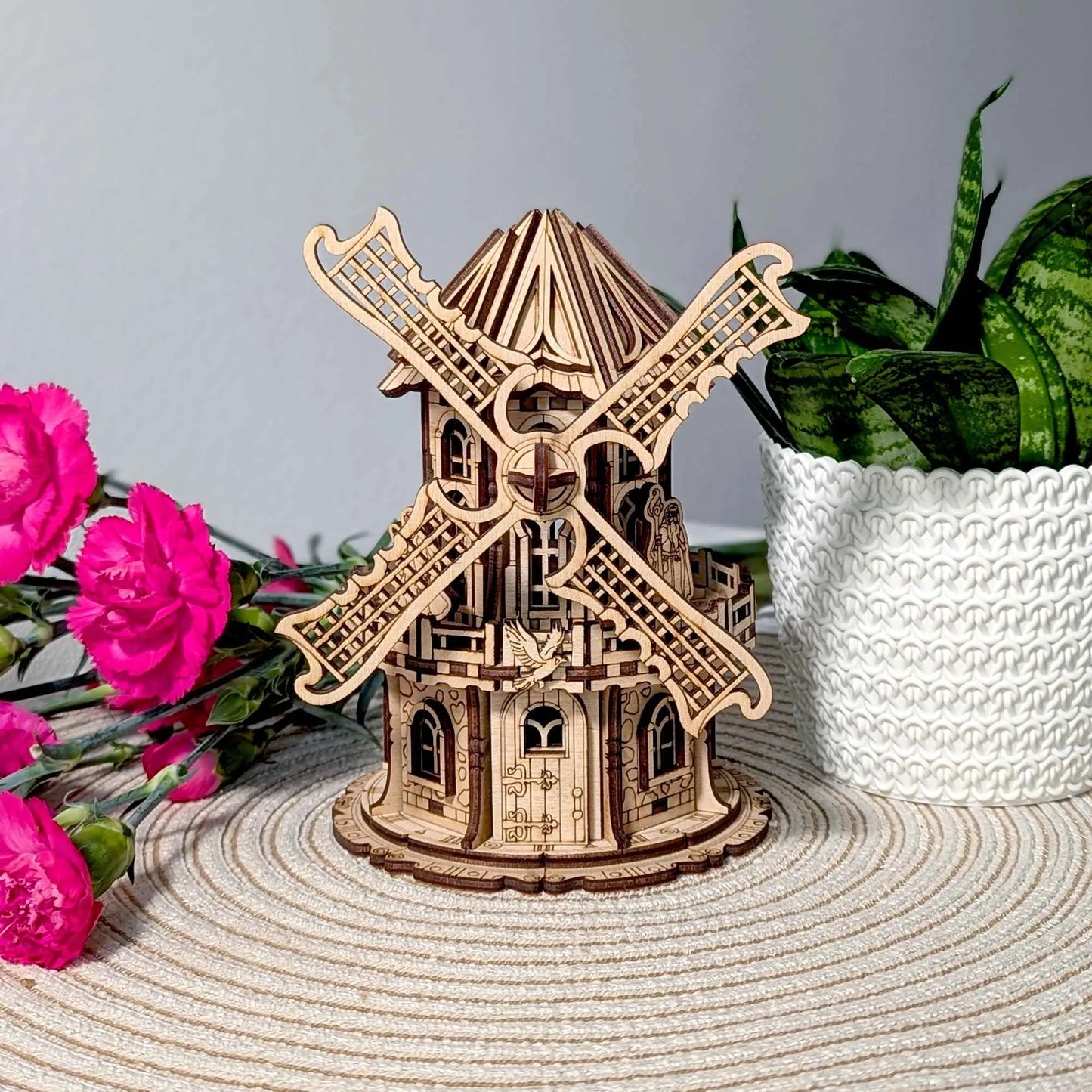 Intricate wooden windmill miniature with detailed laser-cut blades and gothic-style windows, displayed beside bright pink flowers.