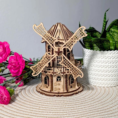 Intricate wooden windmill miniature with detailed laser-cut blades and gothic-style windows, displayed beside bright pink flowers.