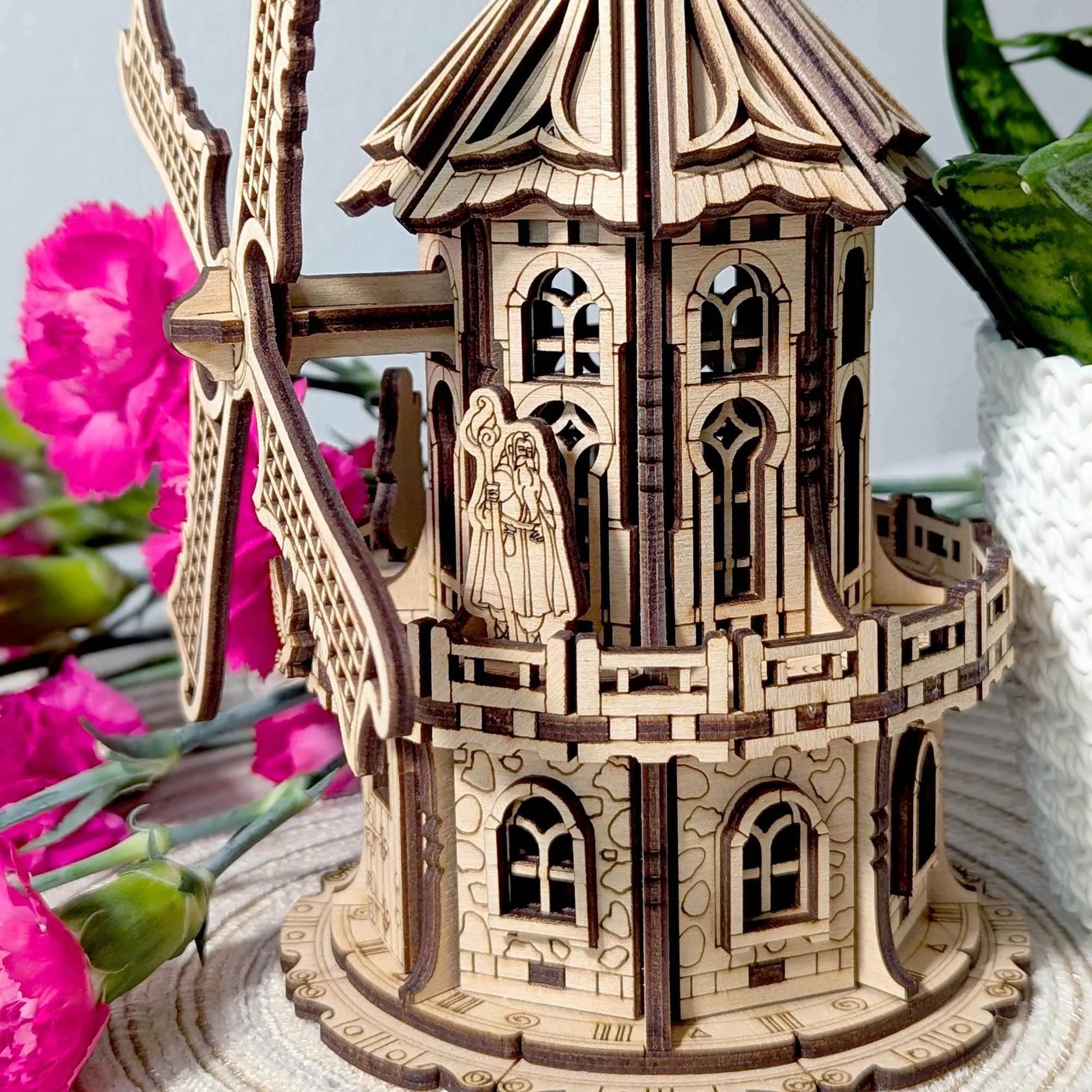 Decorative 3D plywood windmill model with ornate architectural details, photographed on a woven table mat next to indoor plants.