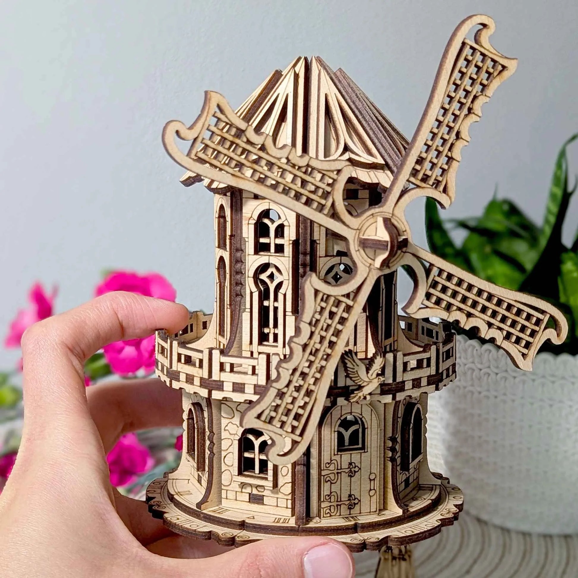 Intricate wooden windmill miniature with detailed laser-cut blades and gothic-style windows, displayed beside bright pink flowers.