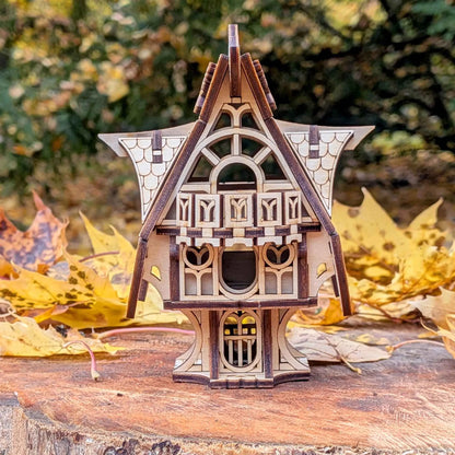 Handmade wooden fantasy cottage model with decorative roof details and arched windows, photographed in an autumn forest setting.