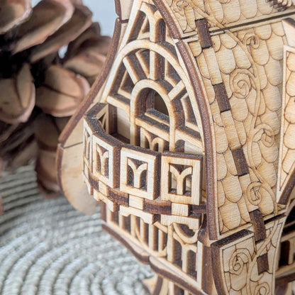 Handmade wooden fantasy cottage model with decorative roof details and arched windows, photographed in an autumn forest setting.