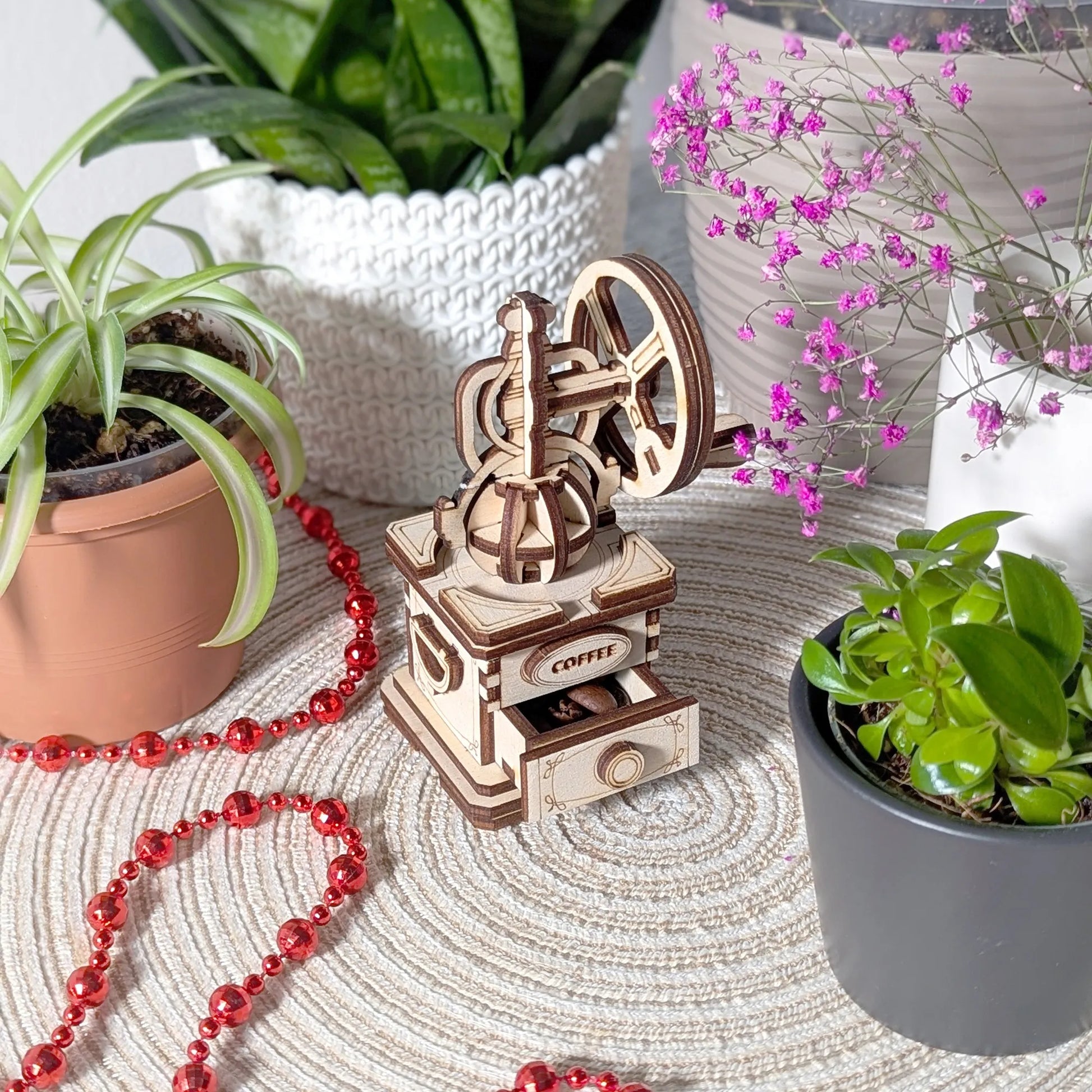 Wooden coffee grinder model on a textured surface with plants and red beads in the background