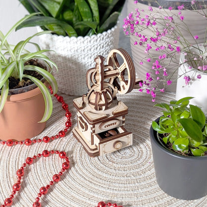Wooden coffee grinder model on a textured surface with plants and red beads in the background